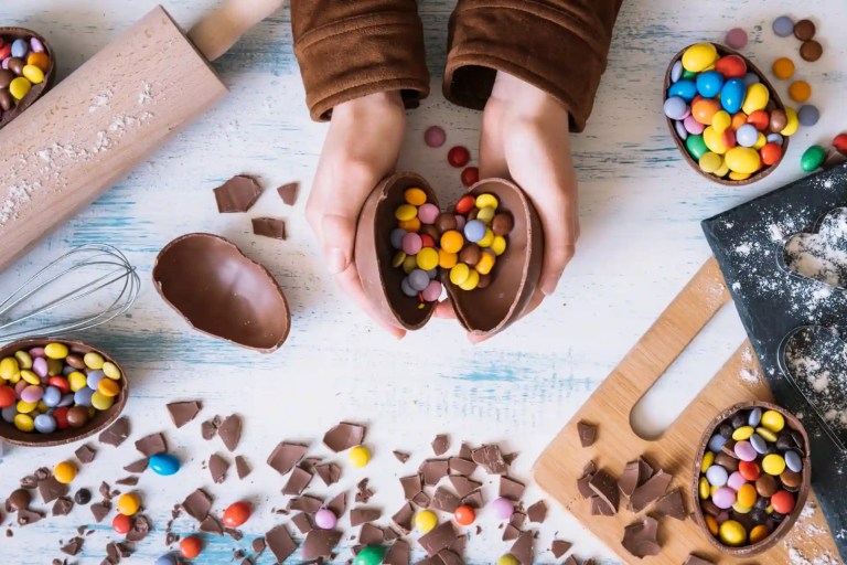 Foto das mãos de uma pessoa segurando as casas de um ovo de chocolate recheado com confetes, em cima de uma mesa com outros ovos de Páscoa coloridos e quitutes.