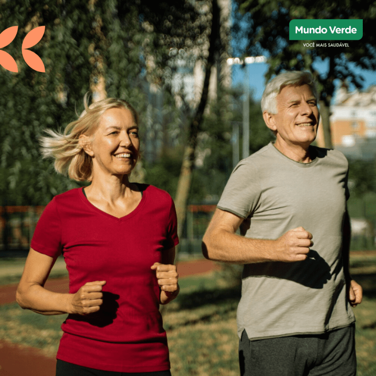 Homem e mulher idosos sorrindo enquanto praticam corrida leve em pista ao ar livre, durante um dia ensolarado, cercados por árvores. A imagem representa um estilo de vida saudável e ativo na terceira idade.