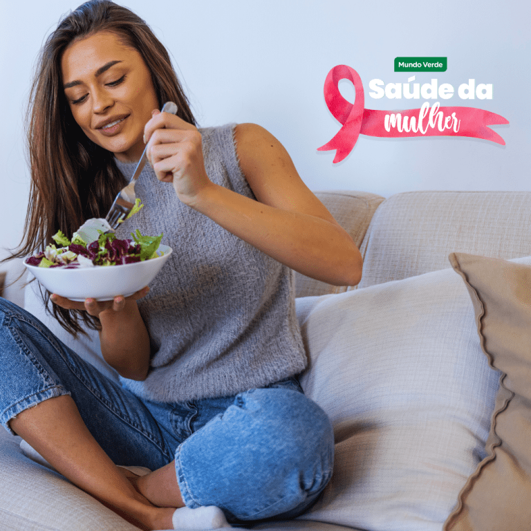 Mulher sorridente comendo salada fresca em casa, representando hábitos alimentares saudáveis que ajudam na prevenção do câncer de mama.