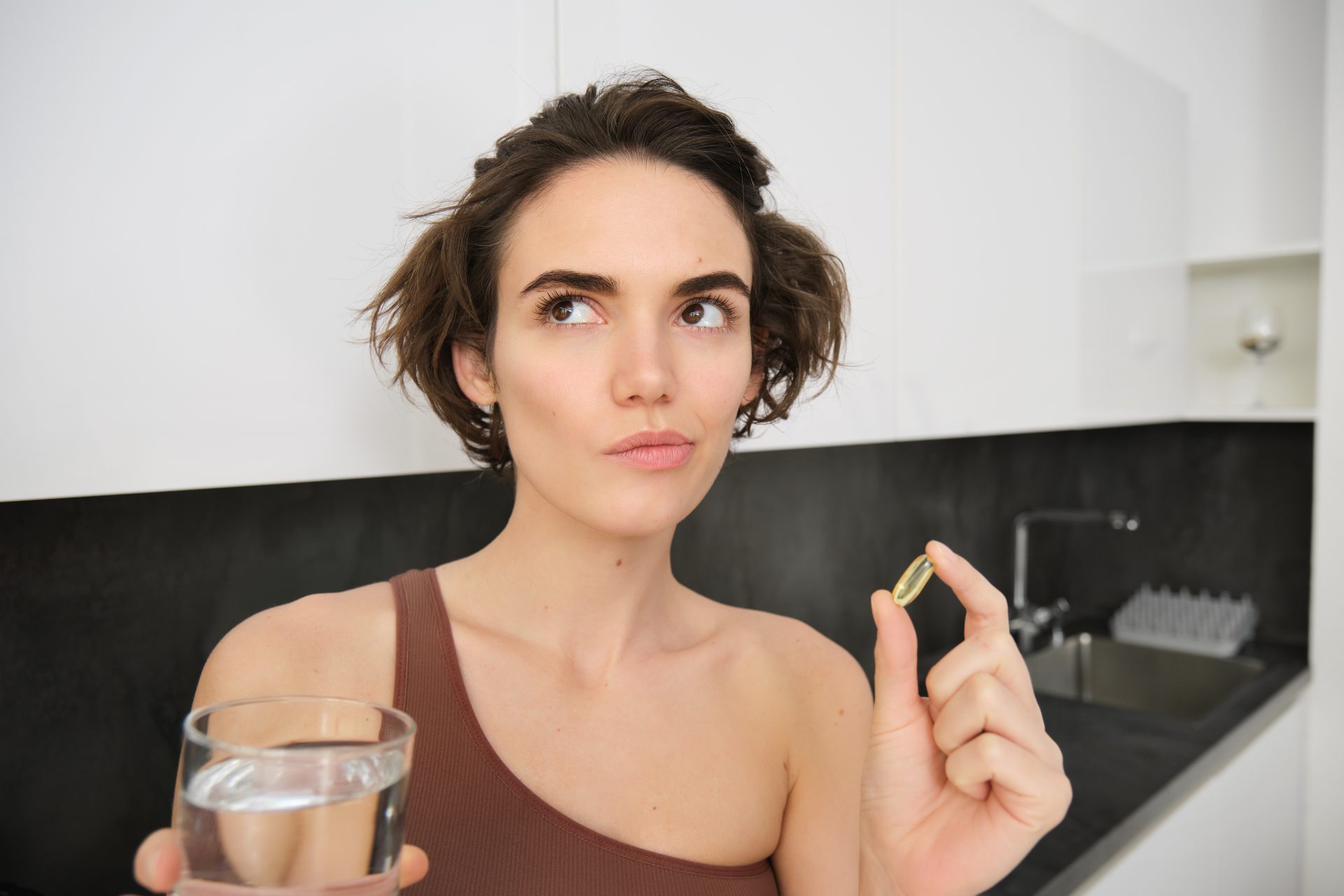 Foto de uma mulher, com expressão de dúvida, segurando um suplemento alimentar em cápsula e um copo de água na cozinha.
