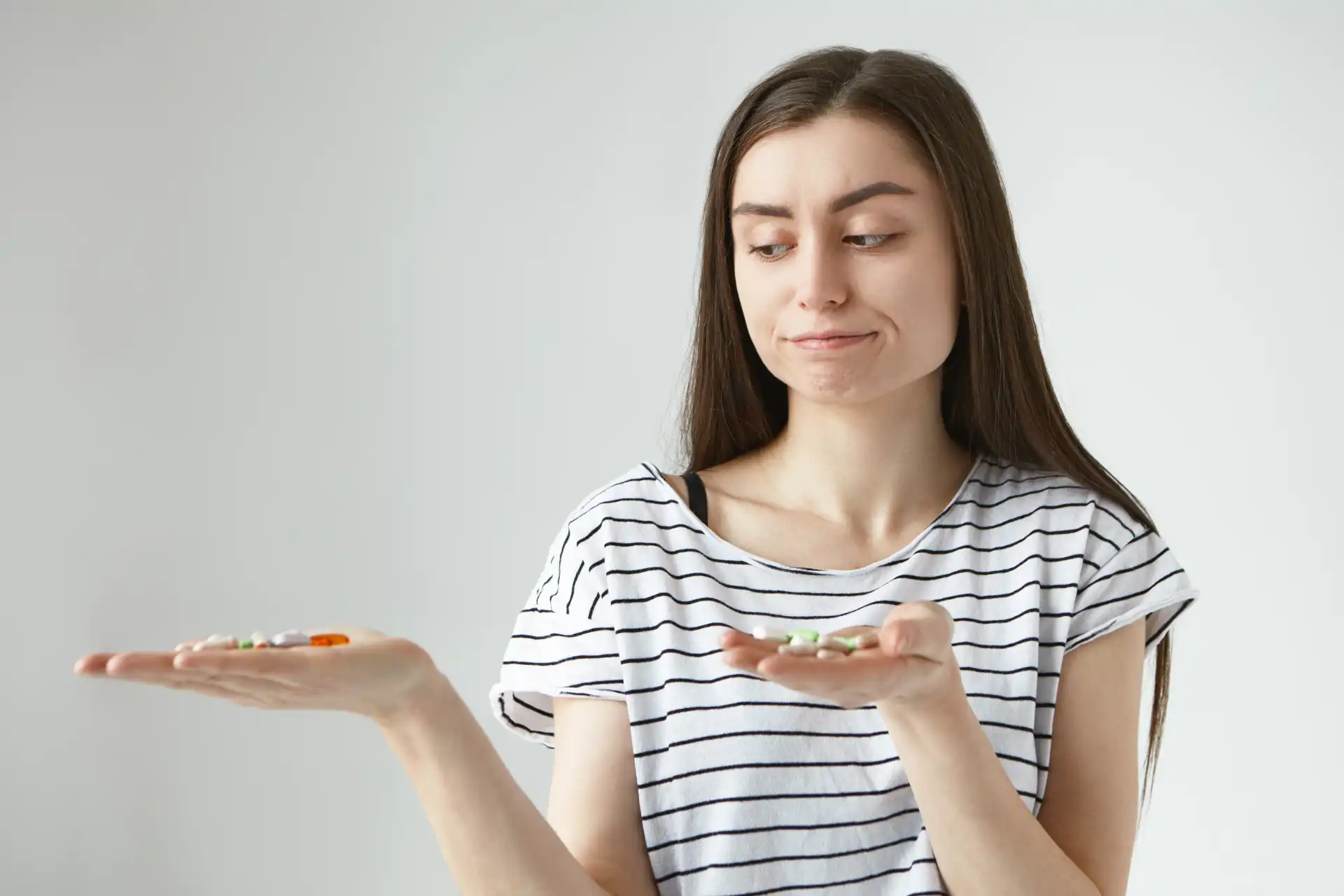 Imagem de uma mulher jovem segurando duas mãos com comprimidos de suplemento alimentar, com expressão de dúvida, vestindo uma camiseta listrada. 