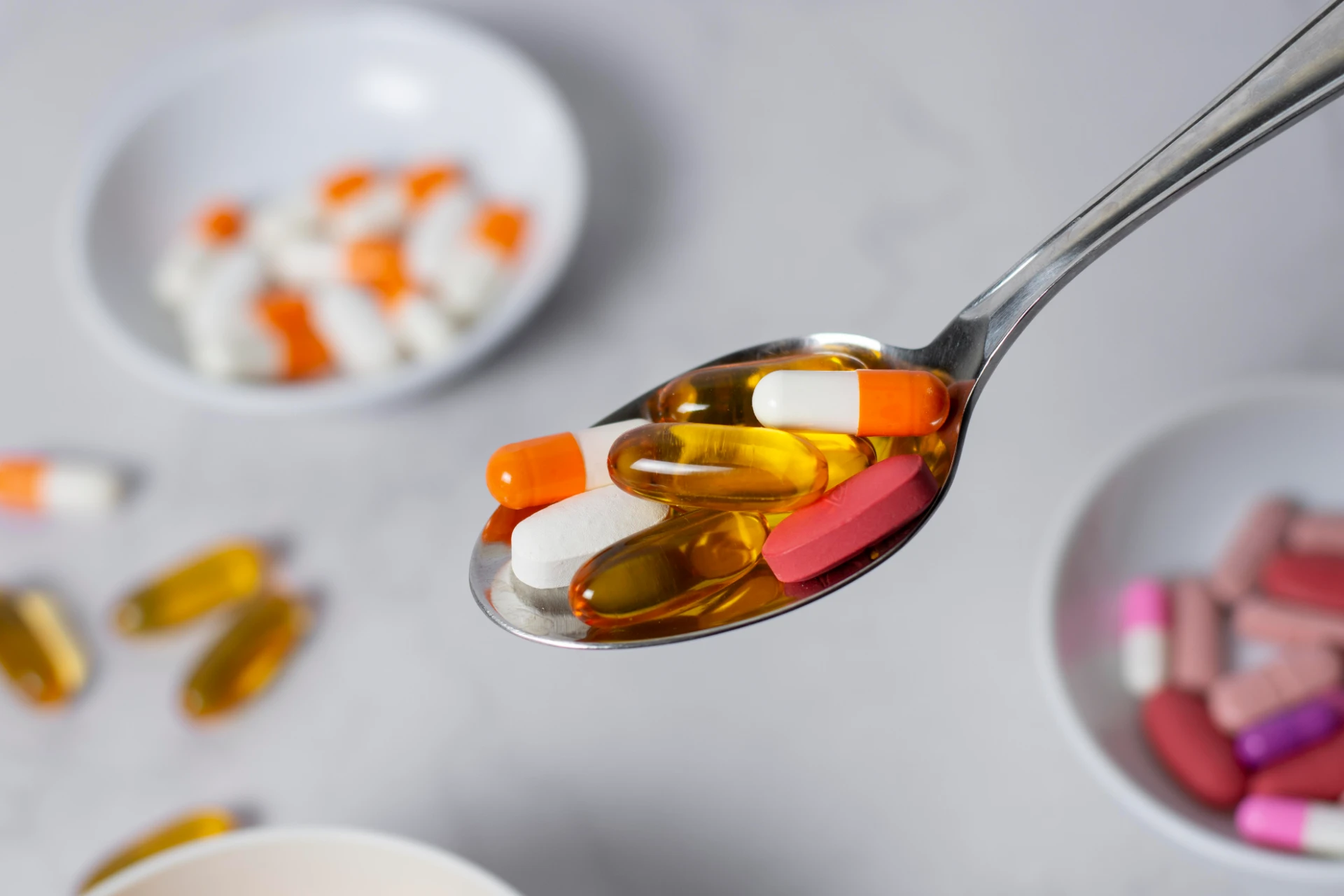Imagem de uma colher com suplementos vitamínicos, vitaminas em cápsulas ou comprimidos diversos, incluindo potes com cápsulas de óleo, comprimidos de cálcio e vitaminas em faixas coloridas. 