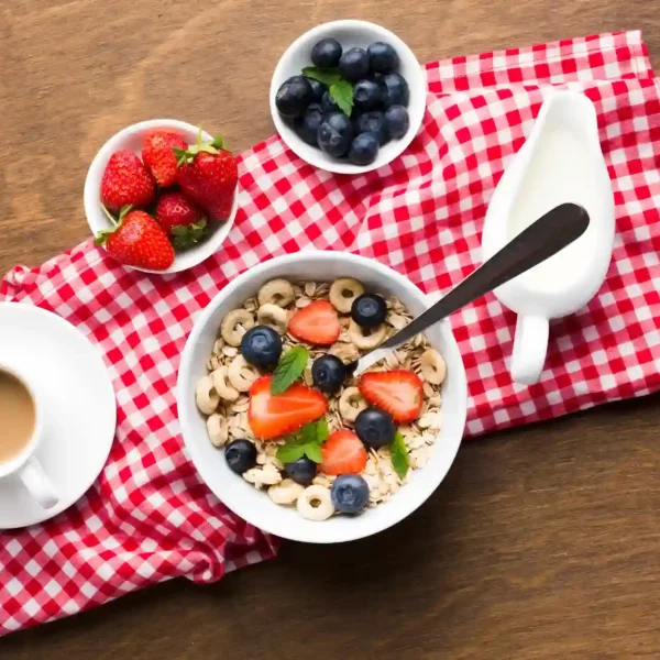 Foto de uma mesa de madeira com alimentos saudáveis para um café da manhã saudável para o Dia das Mães, com uma xícara de café com leite, uma tigela de cereal com frutas, além de dois recipientes com morangos e blueberries.