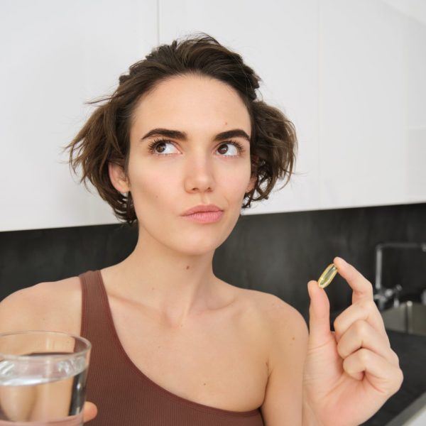 Foto de uma mulher, com expressão de dúvida, segurando um suplemento alimentar em cápsula e um copo de água na cozinha.